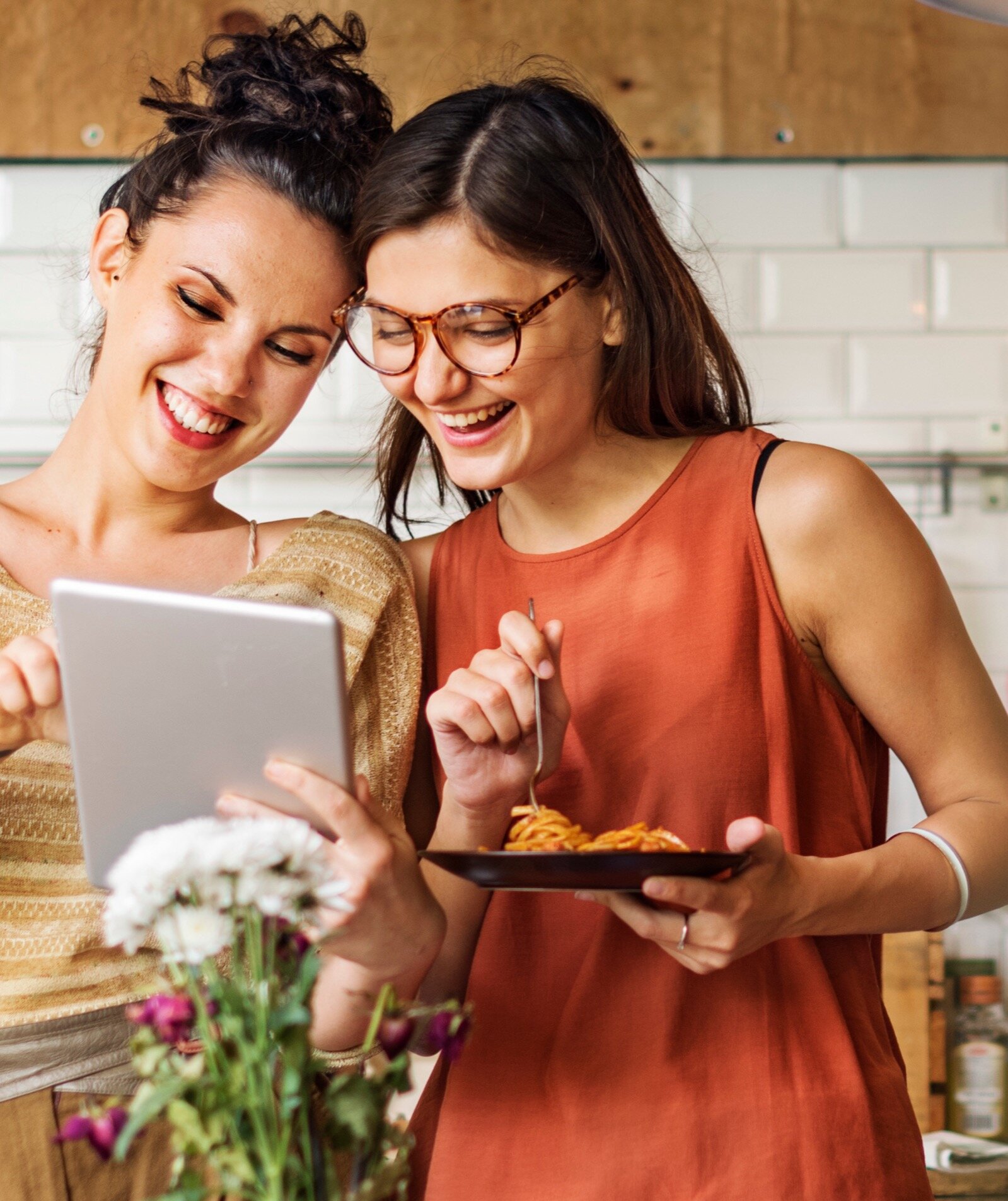 Two women laughing in a kitchen while one eats pasta, a braces-friendly soft food
